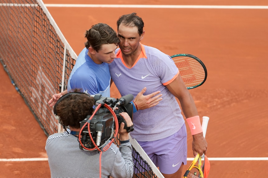 Two tennis players embrace over the net on a red clay surface.