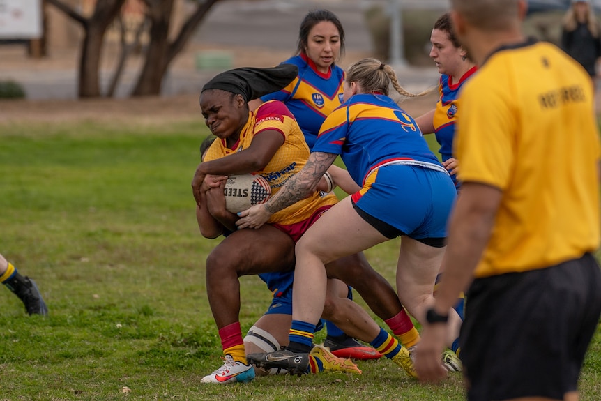 A woman in a yellow jersey is tackled by a woman in a blue jersey, with other players nearby.