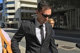 A man in a black suit and tie walks on the footpath holding a woman's hand and wearing sunglasses.