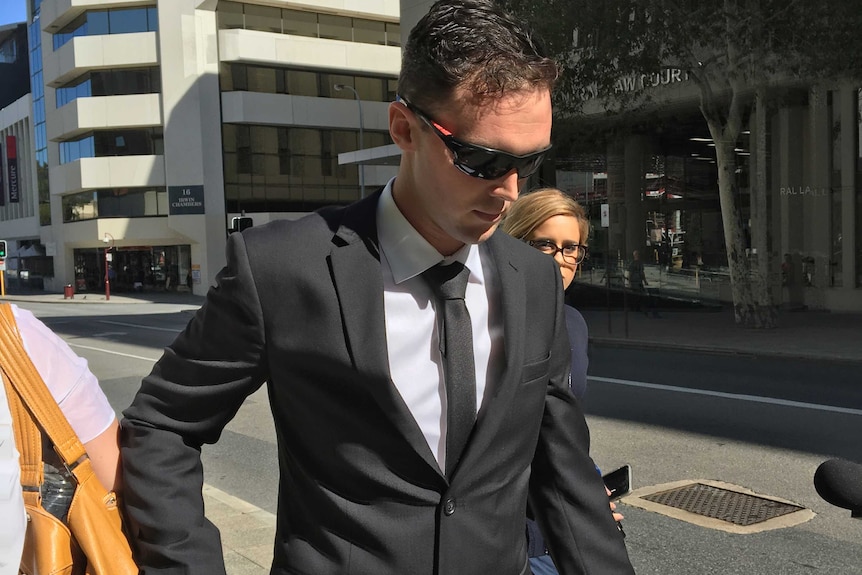 A man in a black suit and tie walks on the footpath holding a woman's hand and wearing sunglasses.