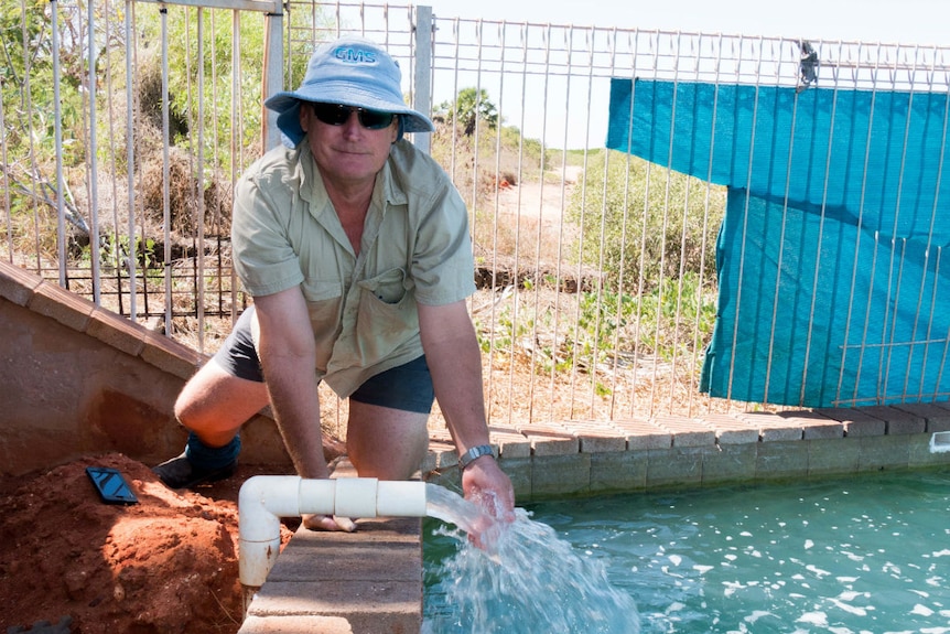 A man in khaki shorts and hat kneels by the side of a pool as a pipe pumps it full of water.