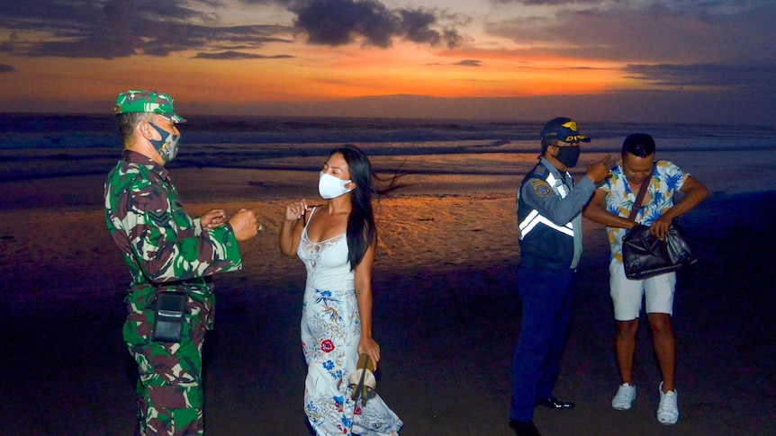 Two tourists in face masks talk to masked soldiers on the beach at sunset