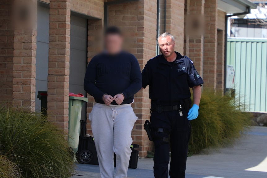 A man wearing track pants is walked from a home with a police officer holding his arm. He is in handcuffs