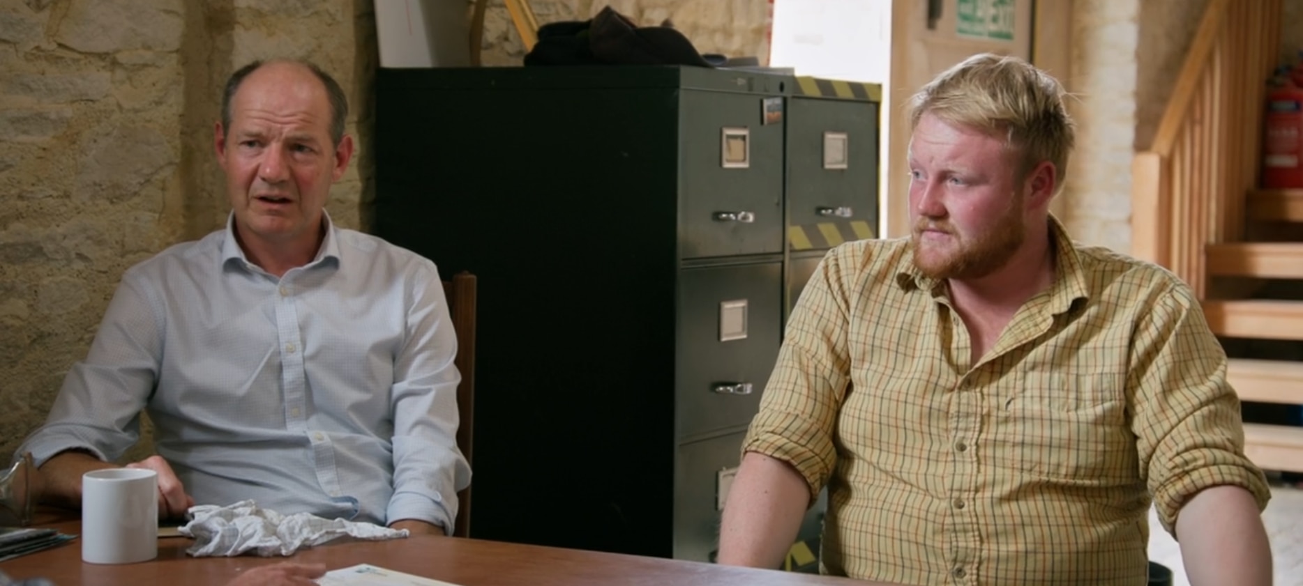 A middle-aged man and a young man look concerned as they sit at a table in a barn.