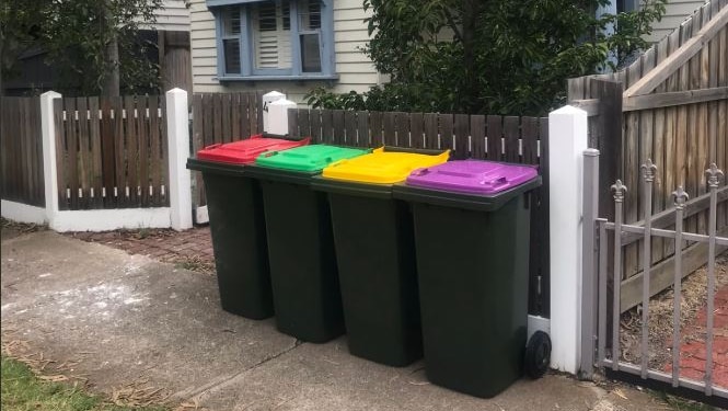 Four bins with red, green, yellow and purple lids sit in a row on the footpath outside a house.