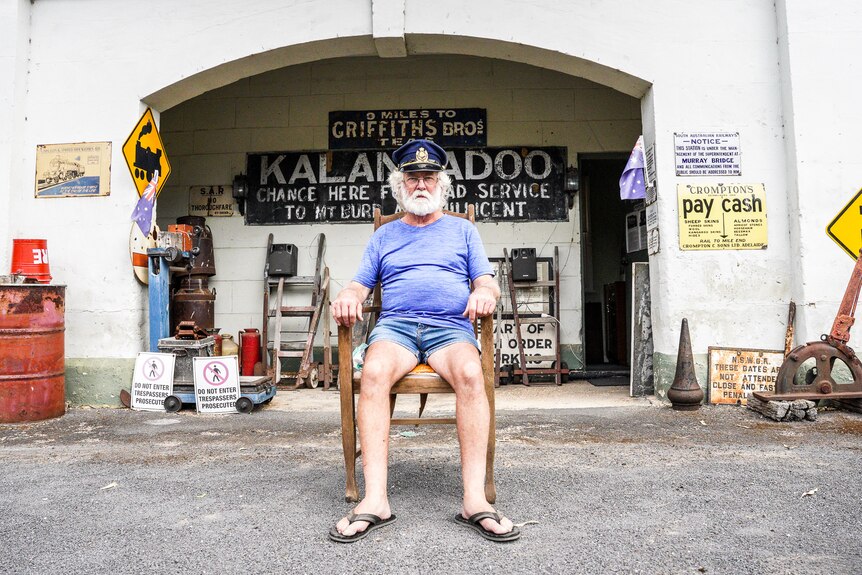 A man with a fluffy white beard sits on a wooden chair outside an old Kalangadoo Railway Station sign.