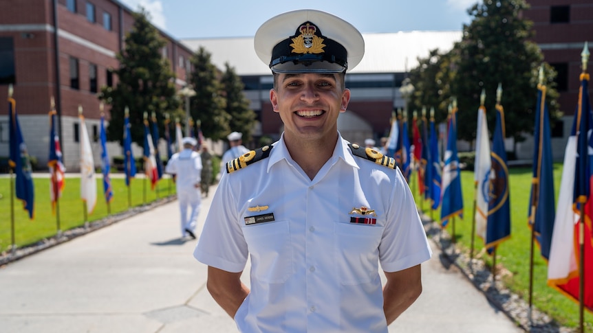 A young man in white naval uniform and peaked hat stands smiling with hands behind his back, outside near flags