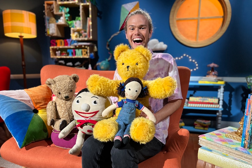 A man sits on the Playschool set with stuffed toys.