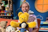 A man sits on the Playschool set with stuffed toys.