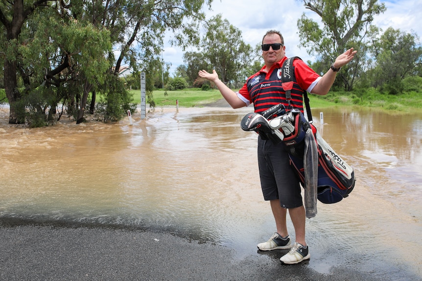 Man with golf clubs stands in front of floodwater.