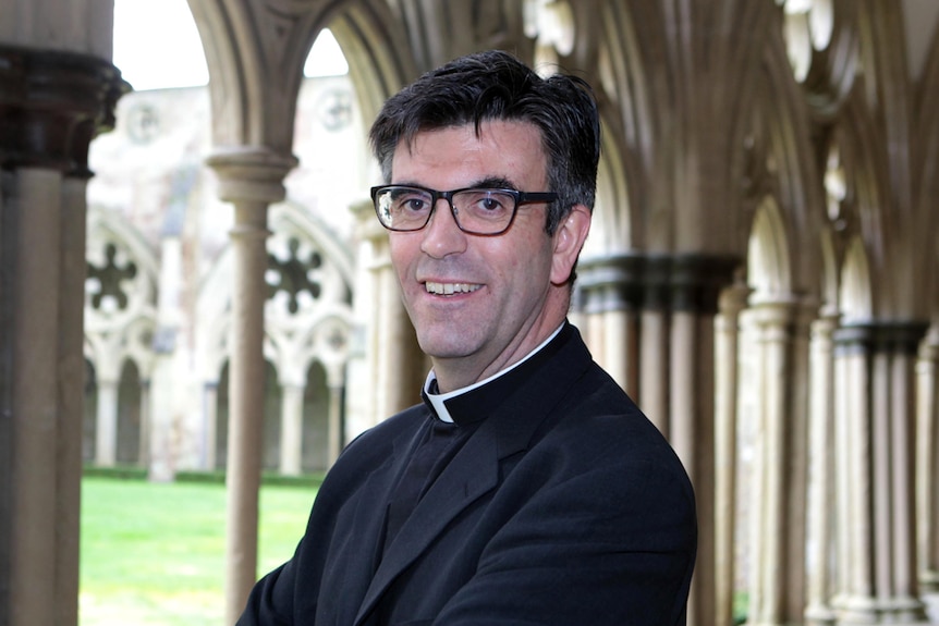 A man with dark hair and glasses, in clerical collar, poses in a church setting