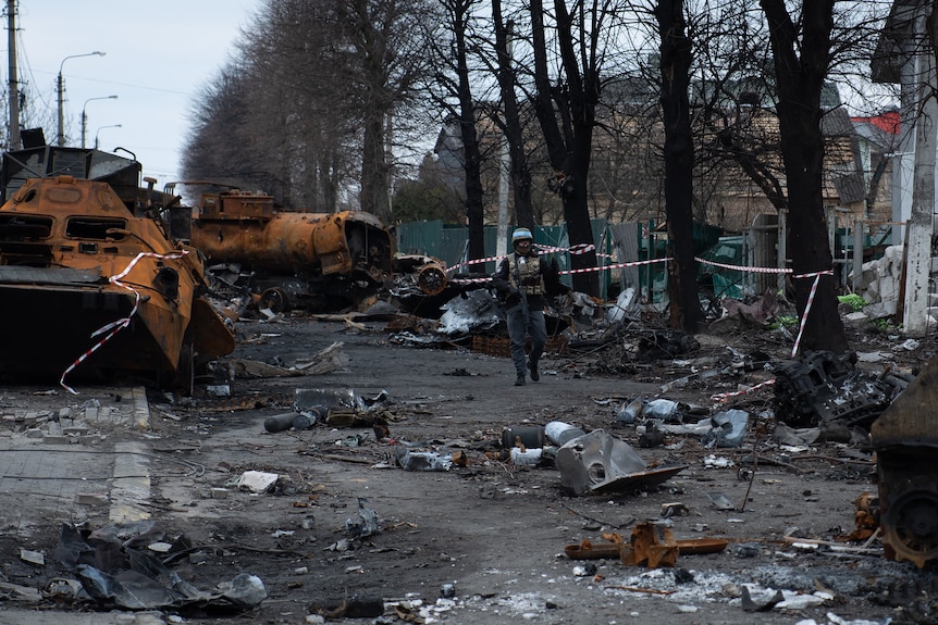 A man wearing a bullet proof vest walks past destroyed vehicles and rubble on a street.