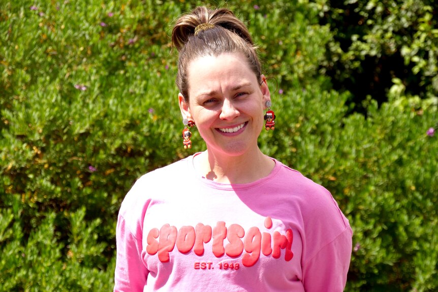 A white woman wearing a pink shirt smiling at the camera with green trees in the background. 