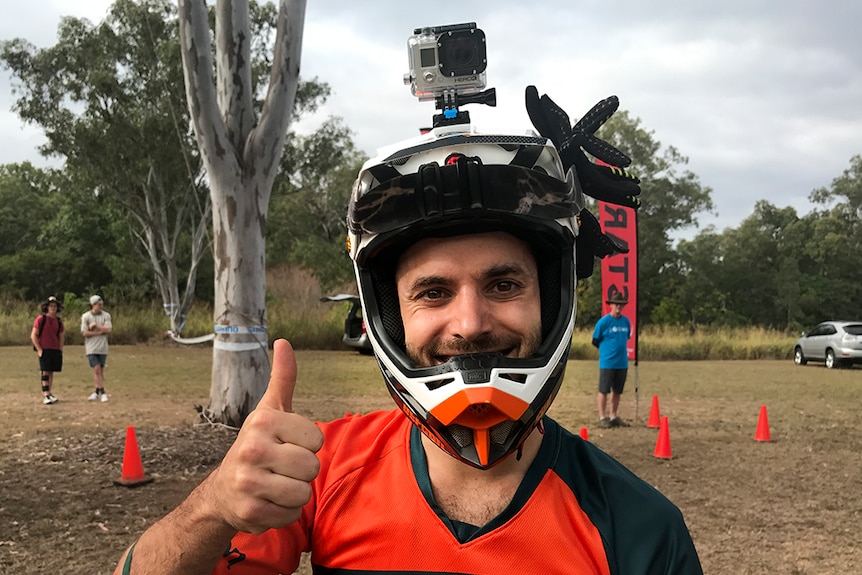 A man with a helmet on and Go Pro on top, smiles with his thumb held up. Start sign in background