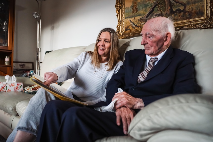 An old man sits on a couch next to a younger woman, looking at a photo album.