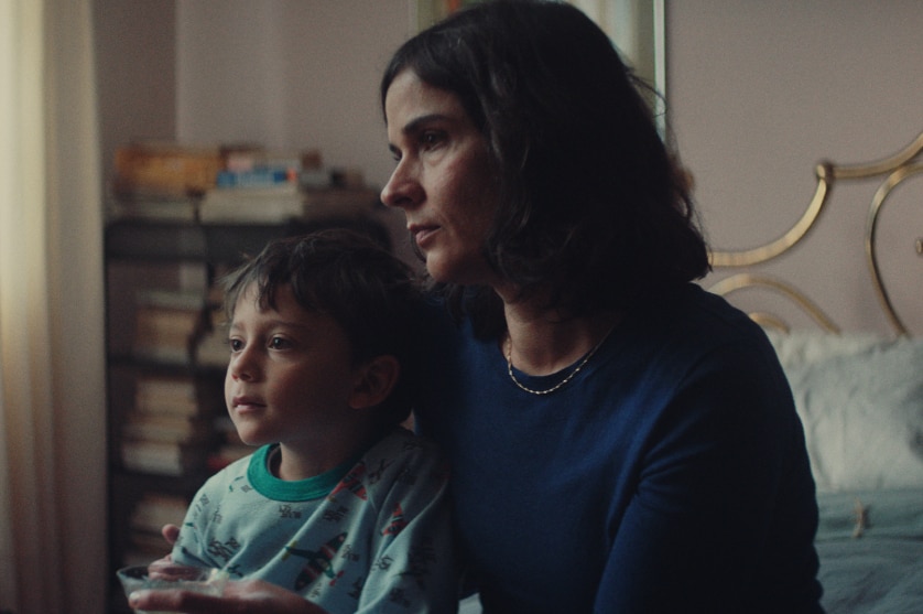 A middle aged woman with shoulder length dark hair and young son sit on bed in bedroom watching something out of frame.