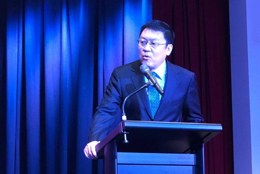 Man in suit stands on stage behind podium with large blue curtain behind him.