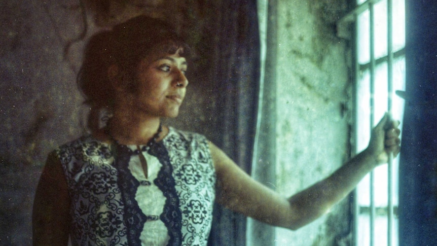 A woman stands in natural light coming from a window, holding onto the window's  protective bars.