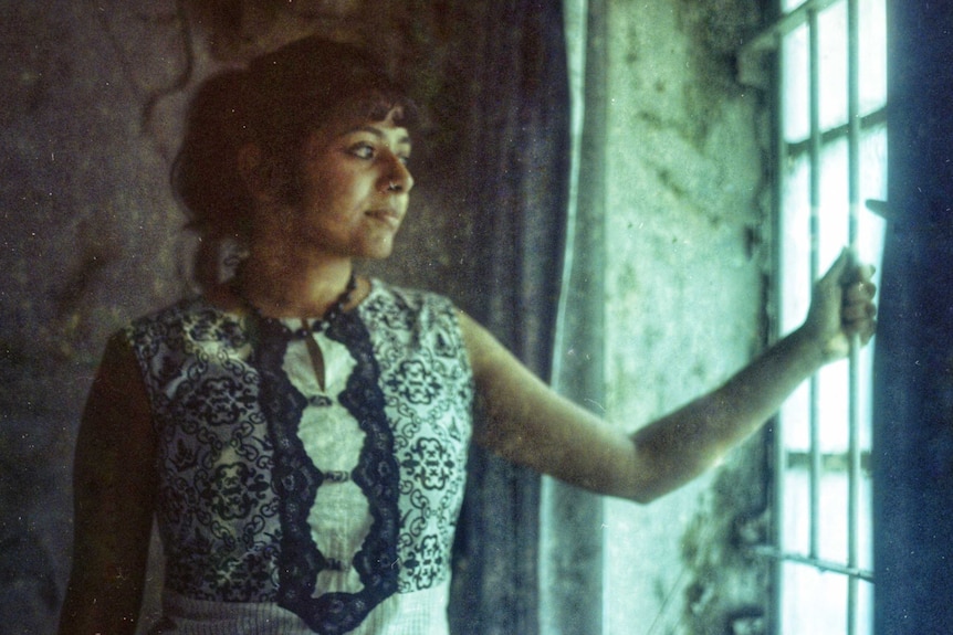 A woman stands in natural light coming from a window, holding onto the window's  protective bars.