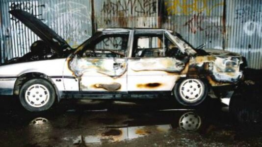A silver car that has been completely damaged by fire parked in front of a shed