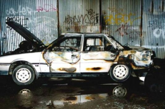 A silver car that has been completely damaged by fire parked in front of a shed