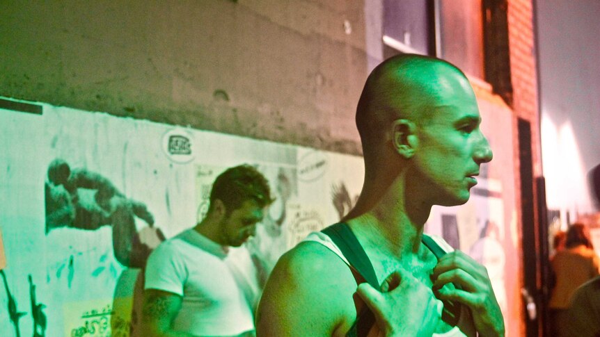 A man in a singlet and backpack with a shaved head stands in an alley