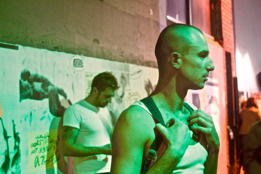 A man in a singlet and backpack with a shaved head stands in an alley