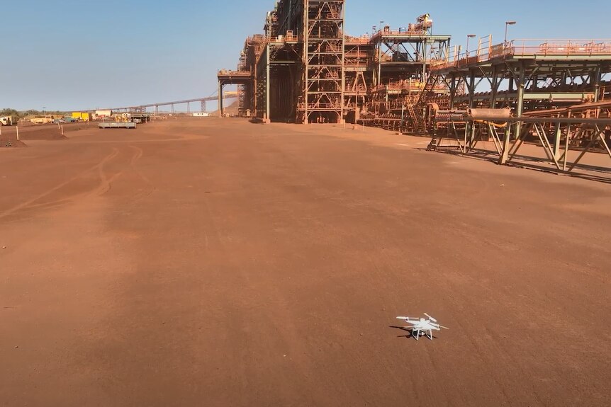 white small-scale drone sits on red dirt in front of mine operations.