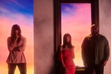 Three members of Khruangbin stand by a large window frame in a wall against a pink/blue sky backdrop