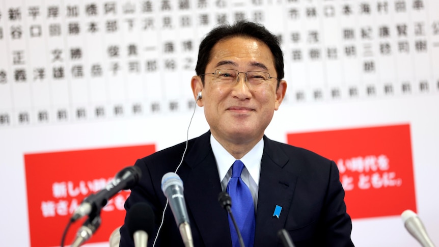 Man in black suit and blue tie smiles in front of microphones