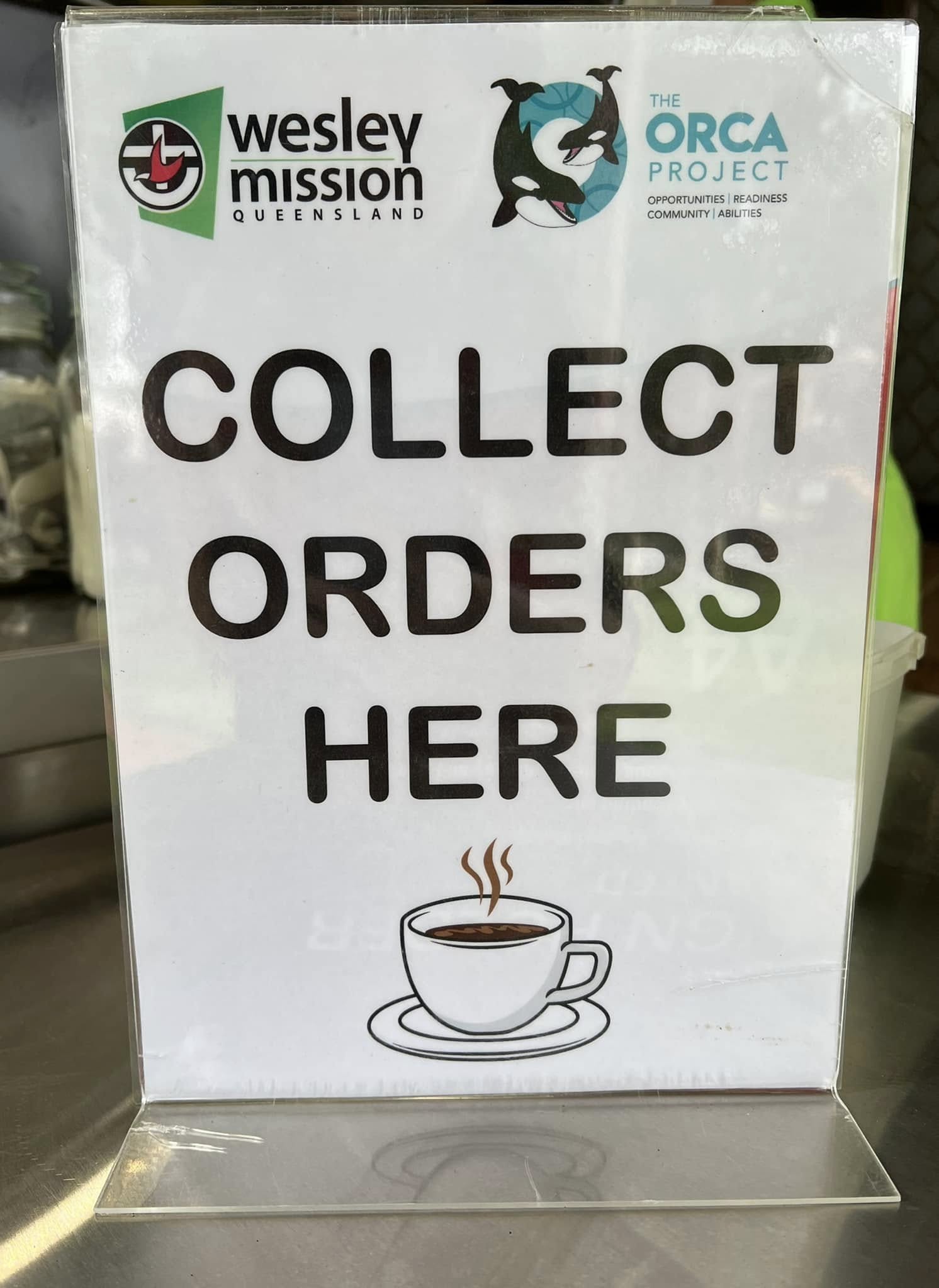 A sign that says, 'Collect orders here'. It also has the ORCA Project and Wesley Mission Queensland logos on it.