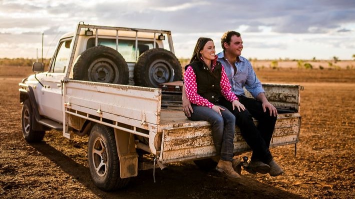 A young couple sit smiling in the back of a white ute in a dry, drought affected paddock