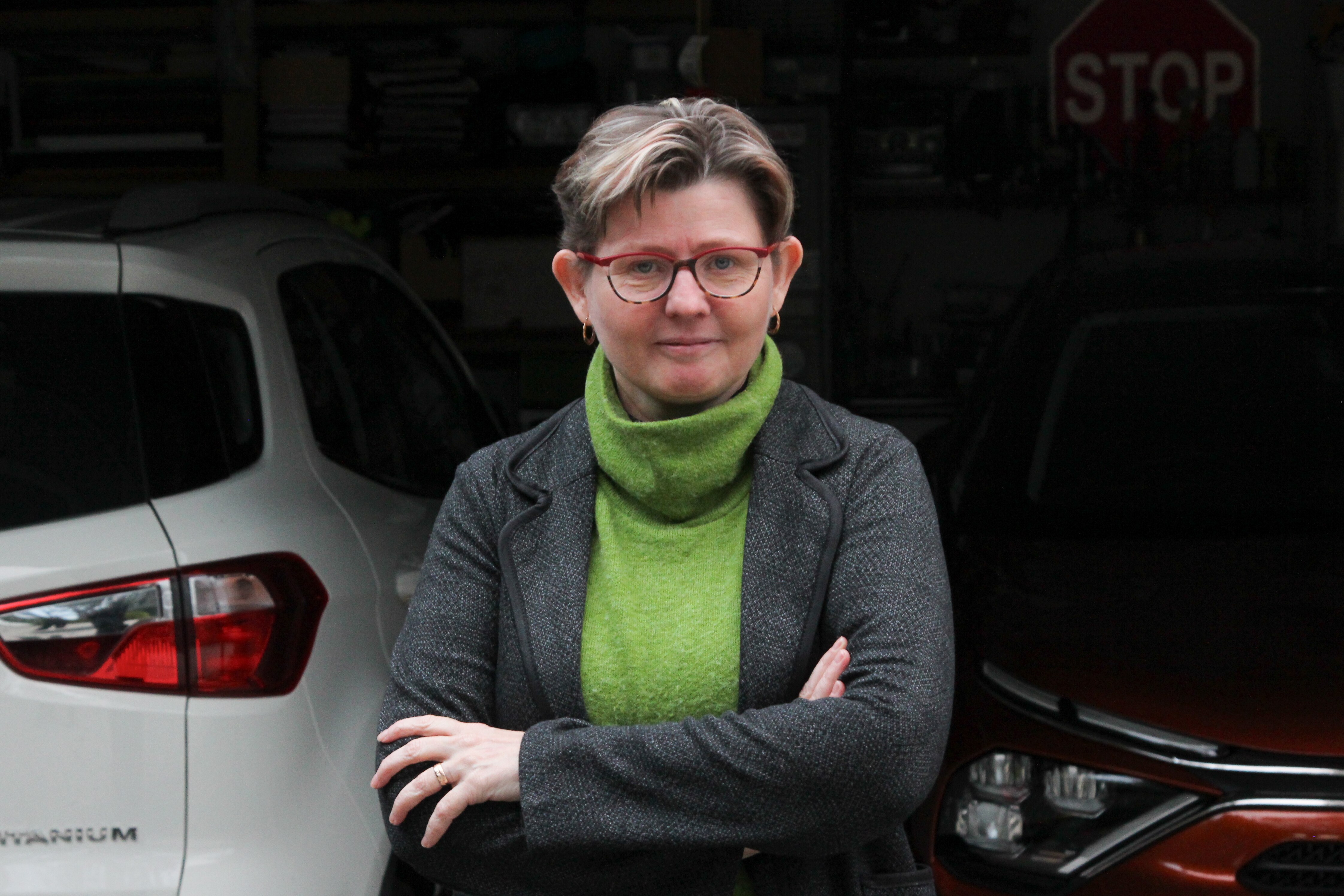 A woman with short hair and a green jumper stands with arms folded in front of a white car and orange car parked in a garage.