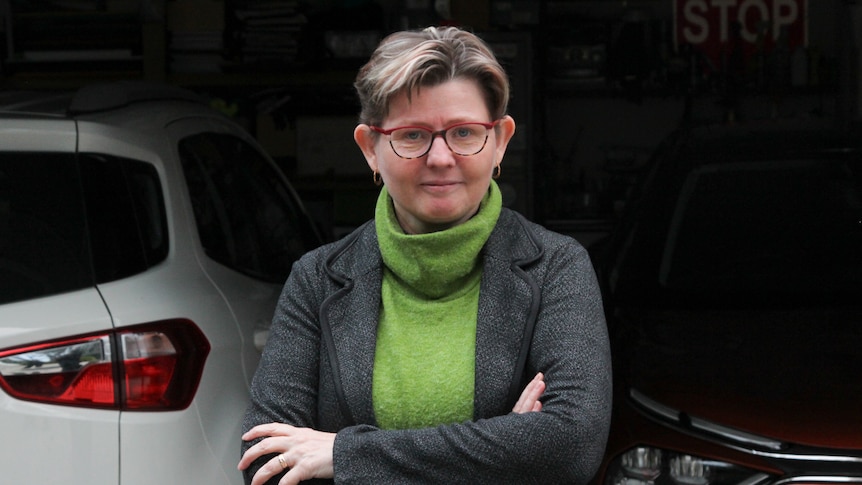 A woman with short hair and a green jumper stands with arms folded in front of a white car and orange car parked in a garage.