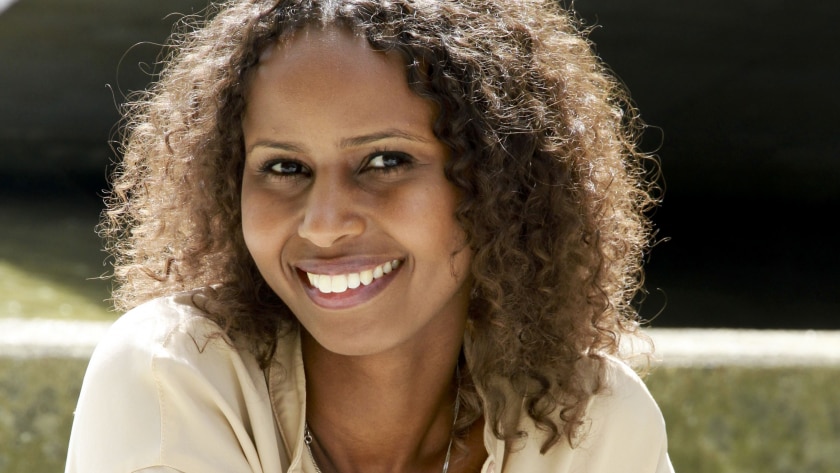Young British-Somali woman with curly shoulder-length brown hair smiling at camera, wearing white top.