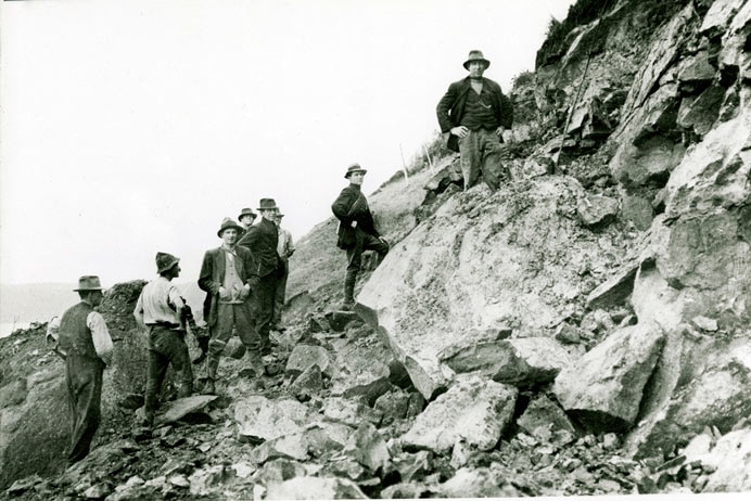 A black and white photo of eight men standing beside large rocks and boulders.