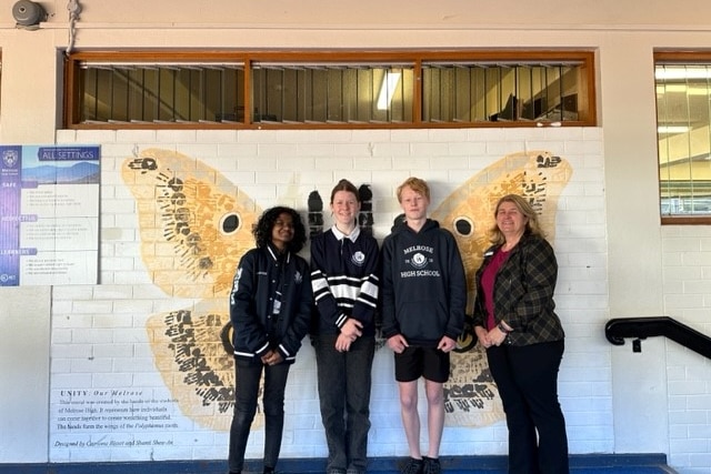 Three students and an adult stand in front of a wall painted with a butterfly.