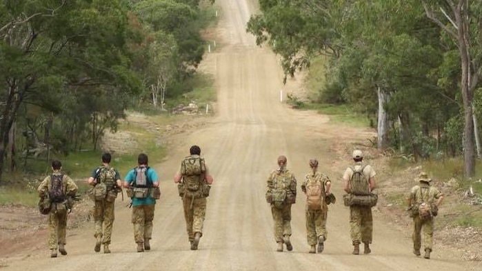 Eight Army people walking along a dirt track surrounded by trees on an overcast day