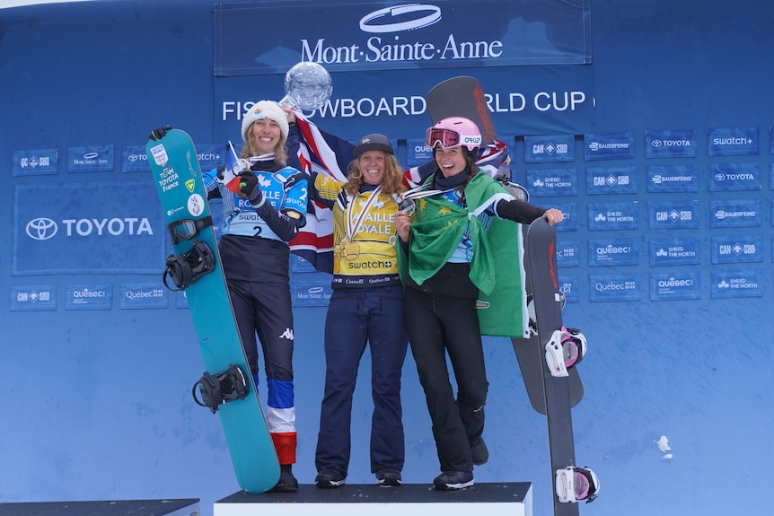 A snowboarder smiles with an Australian flag as she stands on the right of a podium holding a crystal globe trophy.
