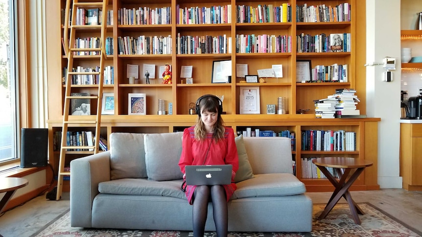 Wearing headphones, Maia Tarrell sits on a couch in front of a bookshelf working at a laptop.