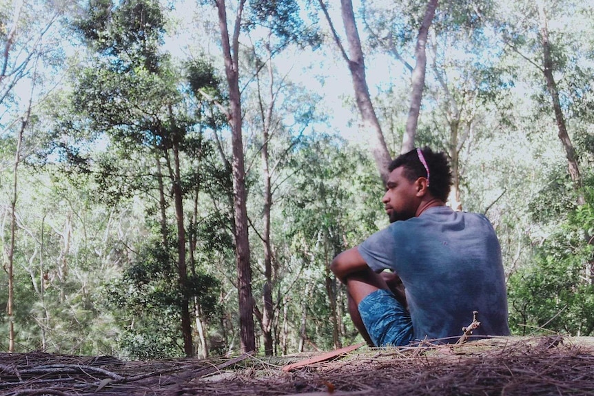 A man staring out into a forest whilst sitting on the ground.