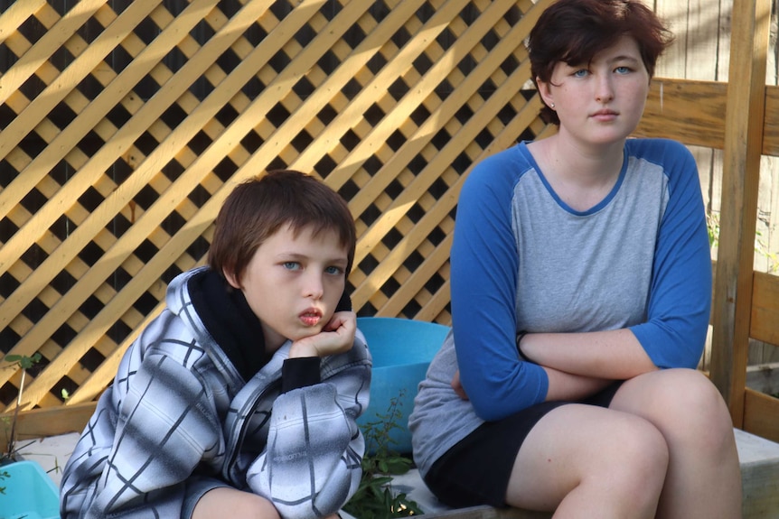 A young boy sits with his hand on his chin while his older sister sits with her arms crossed