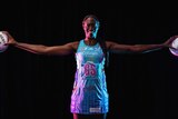Jok wears her light blue and pink Mavericks dress and holds a netball in each hand with her arms stretched out wide