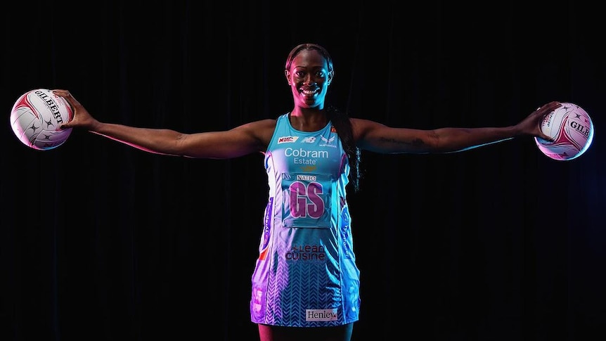 Jok wears her light blue and pink Mavericks dress and holds a netball in each hand with her arms stretched out wide