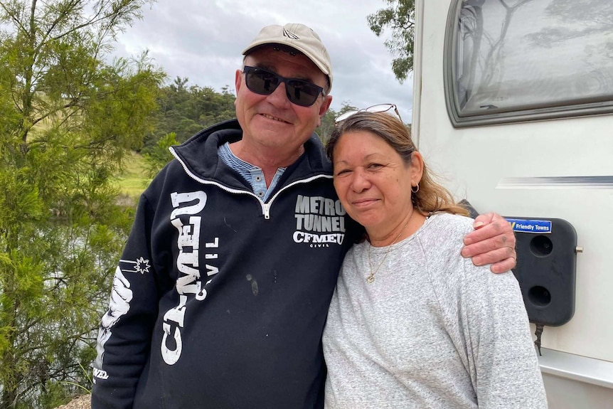 Wayne and Lynne stand in front of their caravan and a river.