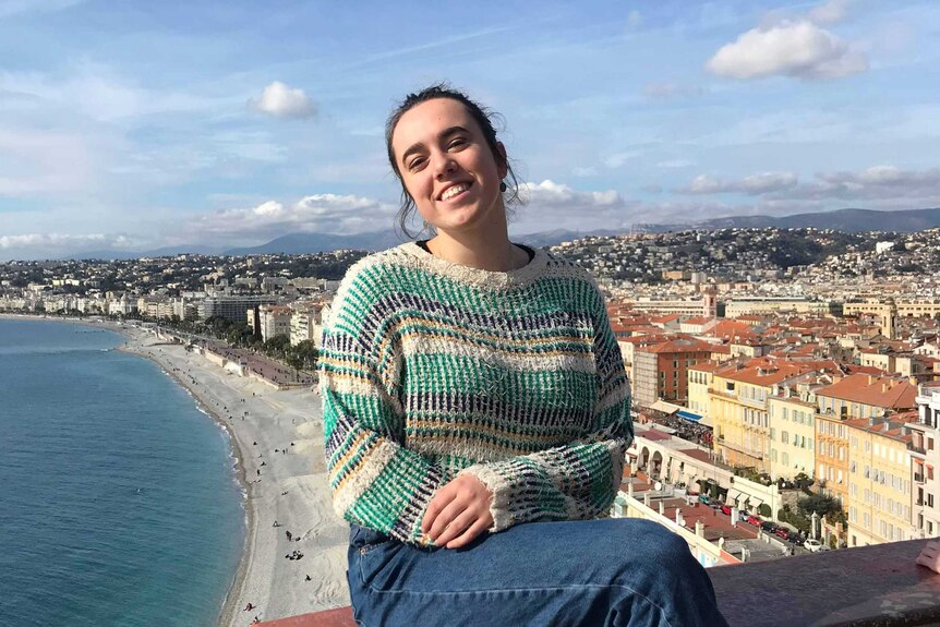 Cassandra Murrell, a young woman, sits and smiles happily in front of the French coastline.
