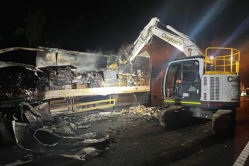 An excavator works on a burnt truck at night.