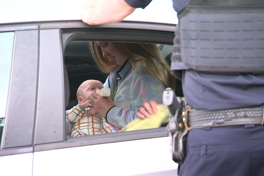 A woman with long blonde hair bottle-feeds her baby in the back of a police car with an office standing outside looking in