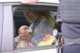 A woman with long blonde hair bottle-feeds her baby in the back of a police car with an office standing outside looking in