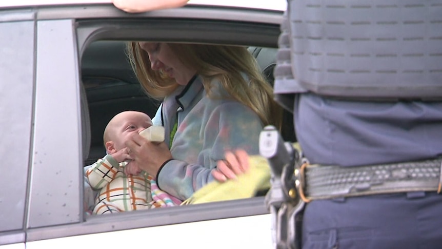 A woman with long blonde hair bottle-feeds her baby in the back of a police car with an office standing outside looking in